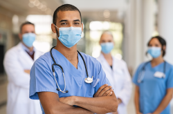 Confident multiethnic male nurse in front of his medical team looking at camera wearing face mask during covid-19 outbreak. Happy and proud indian young surgeon standing in front of his colleagues wearing surgical mask for prevention against coronavirus. Portrait of mixed race doctor with medical staff in background at hospital.